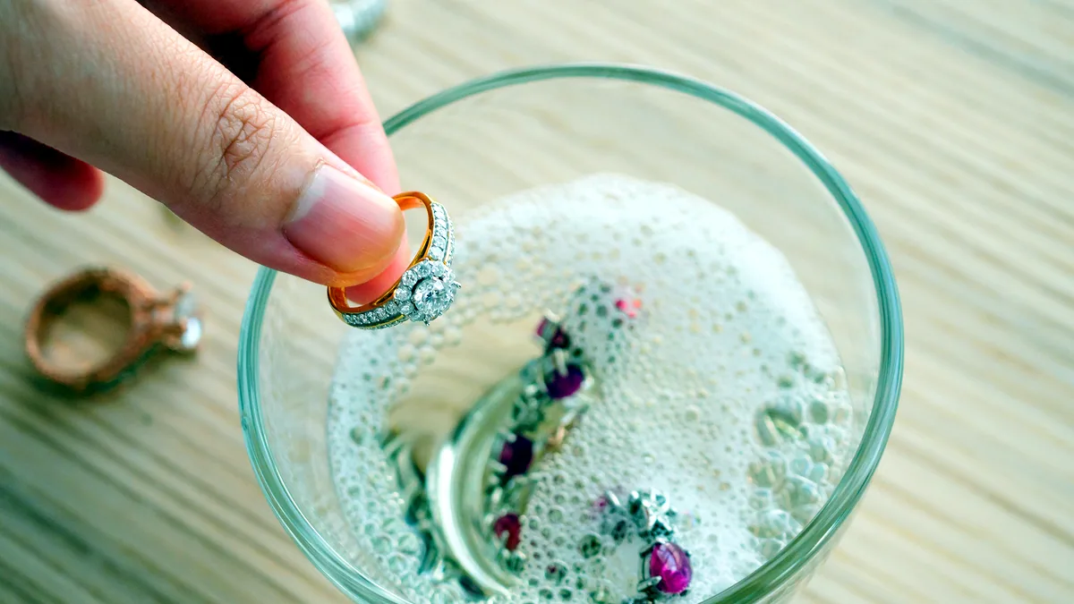 A silver ring submerged in a small glass bowl of soapy water, bubbles clinging to its surface under cool light.
