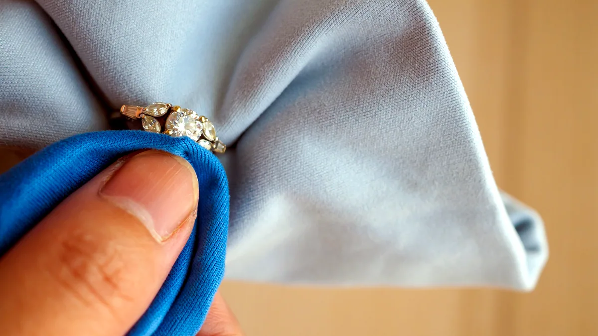 A silver ring resting on a folded dark polishing cloth, a small soft-bristle brush lying beside it on charcoal.